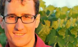 Man with glasses smiling outdoors near leaves.