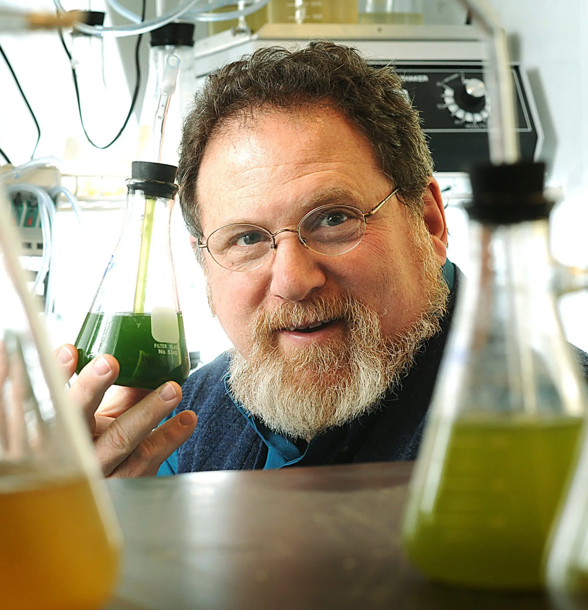 A man with glasses and a beard smiles behind laboratory glassware.