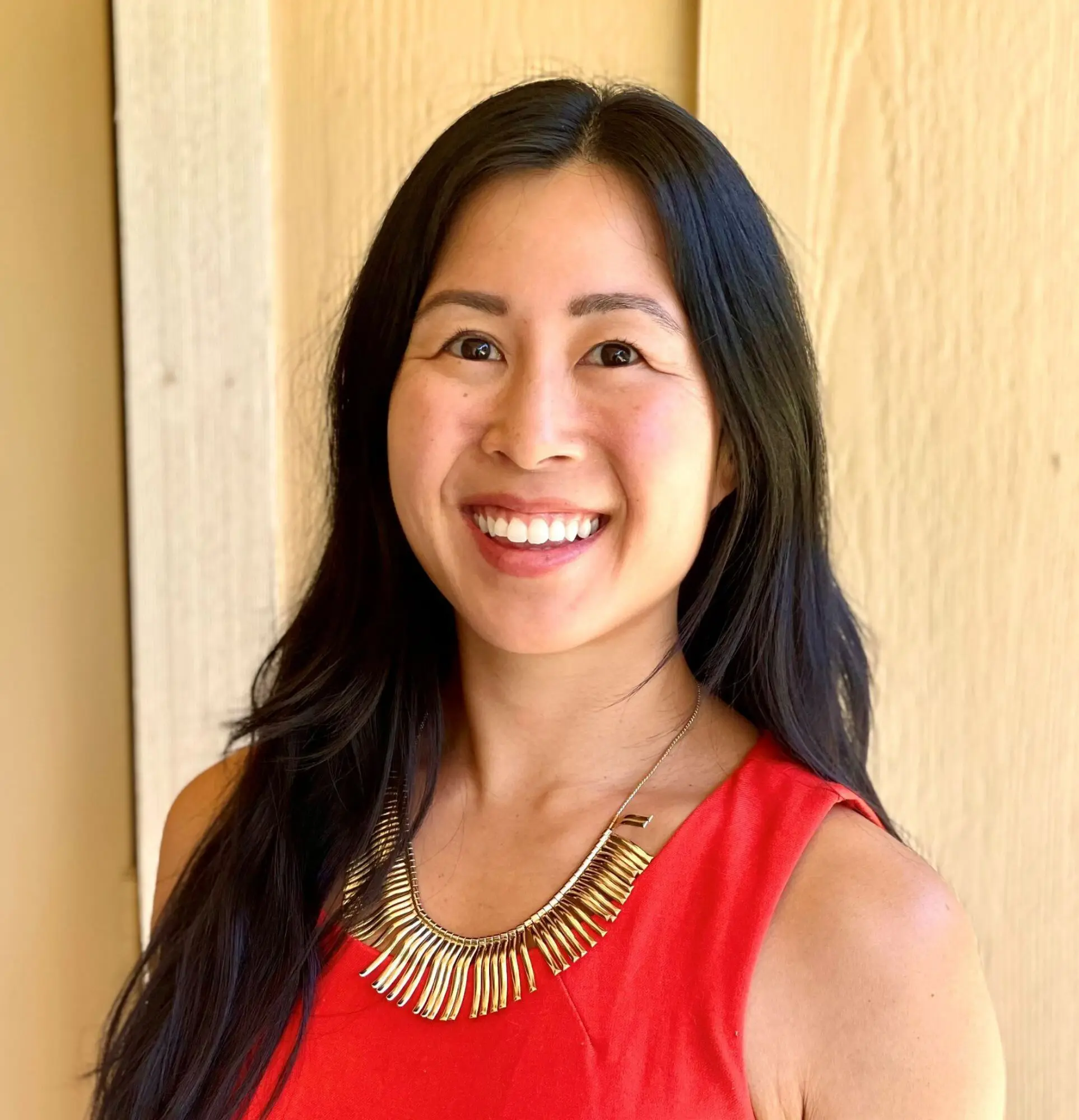 Smiling woman with long black hair wearing a red top and statement necklace.