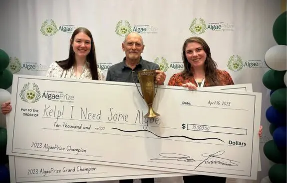 Three people holding a giant check and a trophy, celebrating a win.