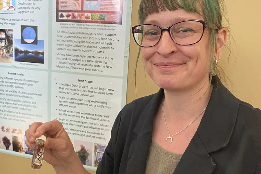 Smiling woman holding a small object near a scientific poster.