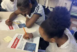 Two girls focused on writing and reading a newspaper article together.