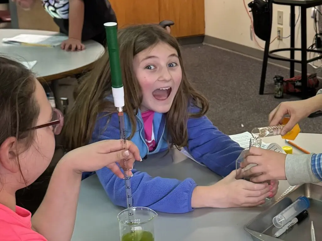 Excited girl in a science class holding a test tube setup.