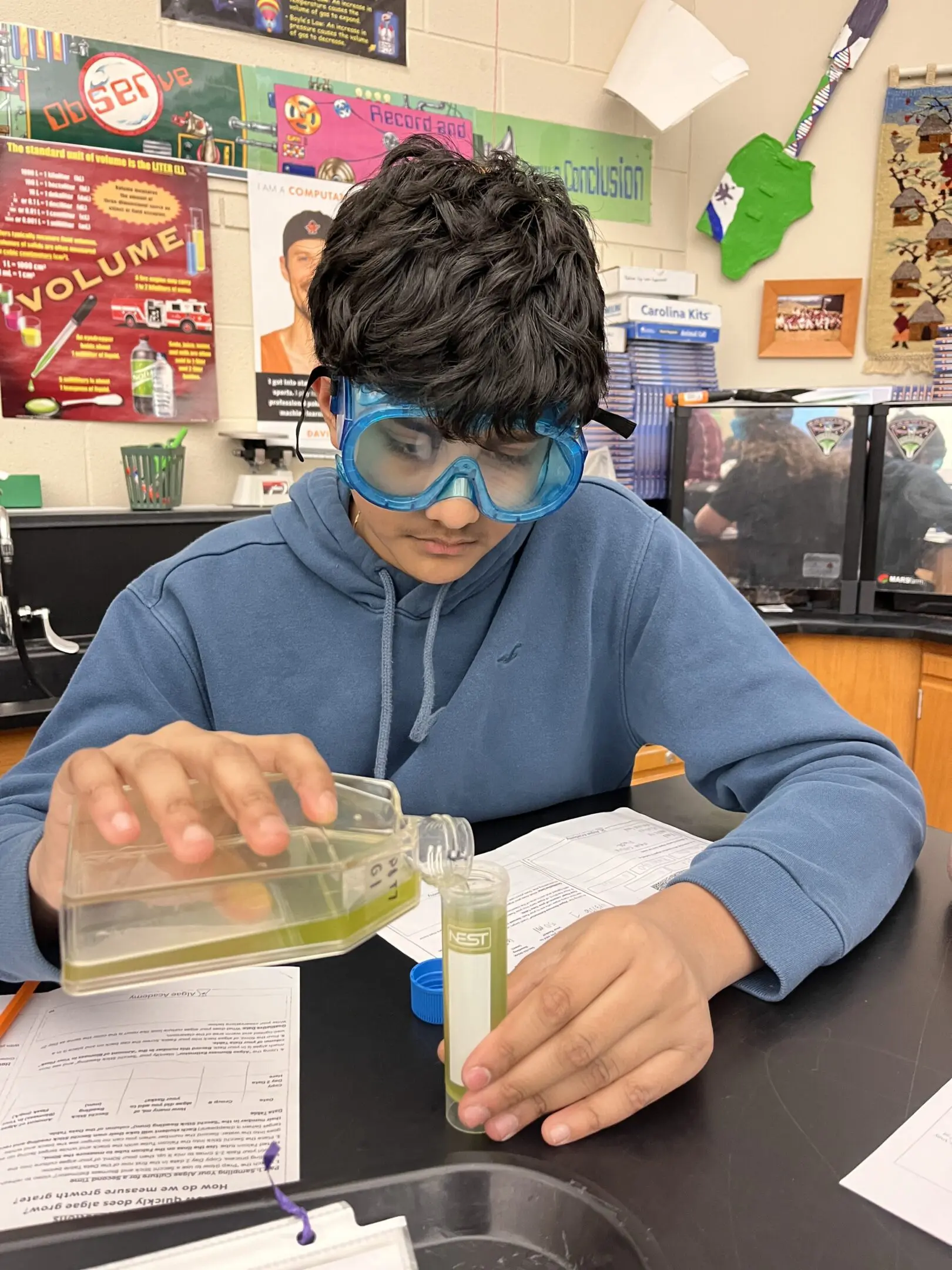 Student wearing goggles pouring liquid into a test tube in a lab.