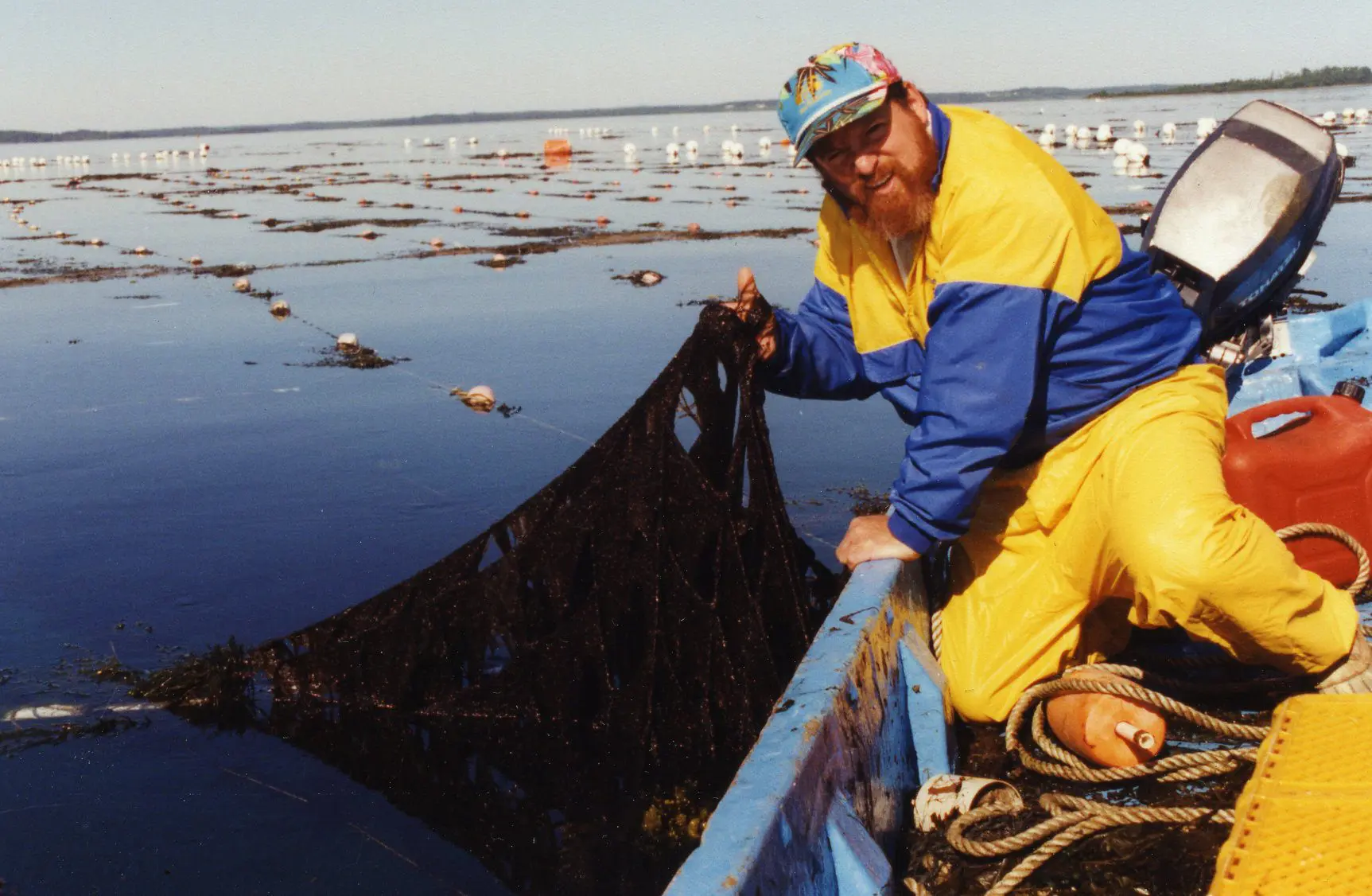 A fisherman in yellow and blue gear holds a net on a boat with floating debris around.