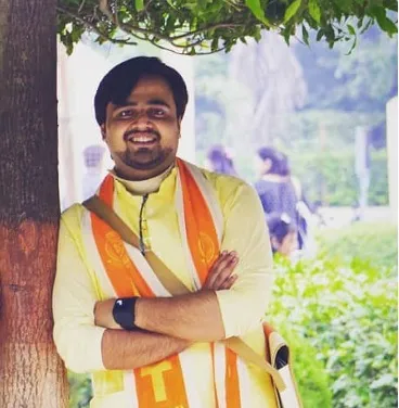 Man smiling outdoors with arms crossed under a tree.