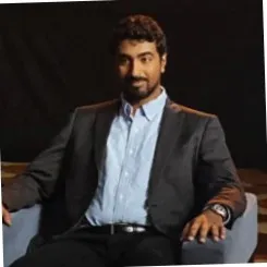 Man in a dark suit sitting on a chair with a confident expression.